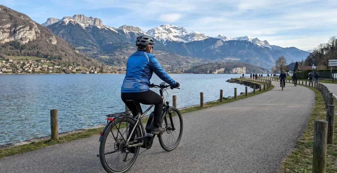 Un cycliste de dos pédale sur un vélo à assistance électrique le long d'une piste cyclable bordant le lac d'Annecy, avec les montagnes des Alpes en arrière-plan sous une lumière matinale douce