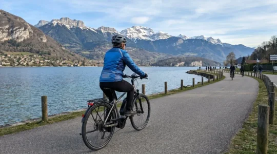 Un cycliste de dos pédale sur un vélo à assistance électrique le long d'une piste cyclable bordant le lac d'Annecy, avec les montagnes des Alpes en arrière-plan sous une lumière matinale douce