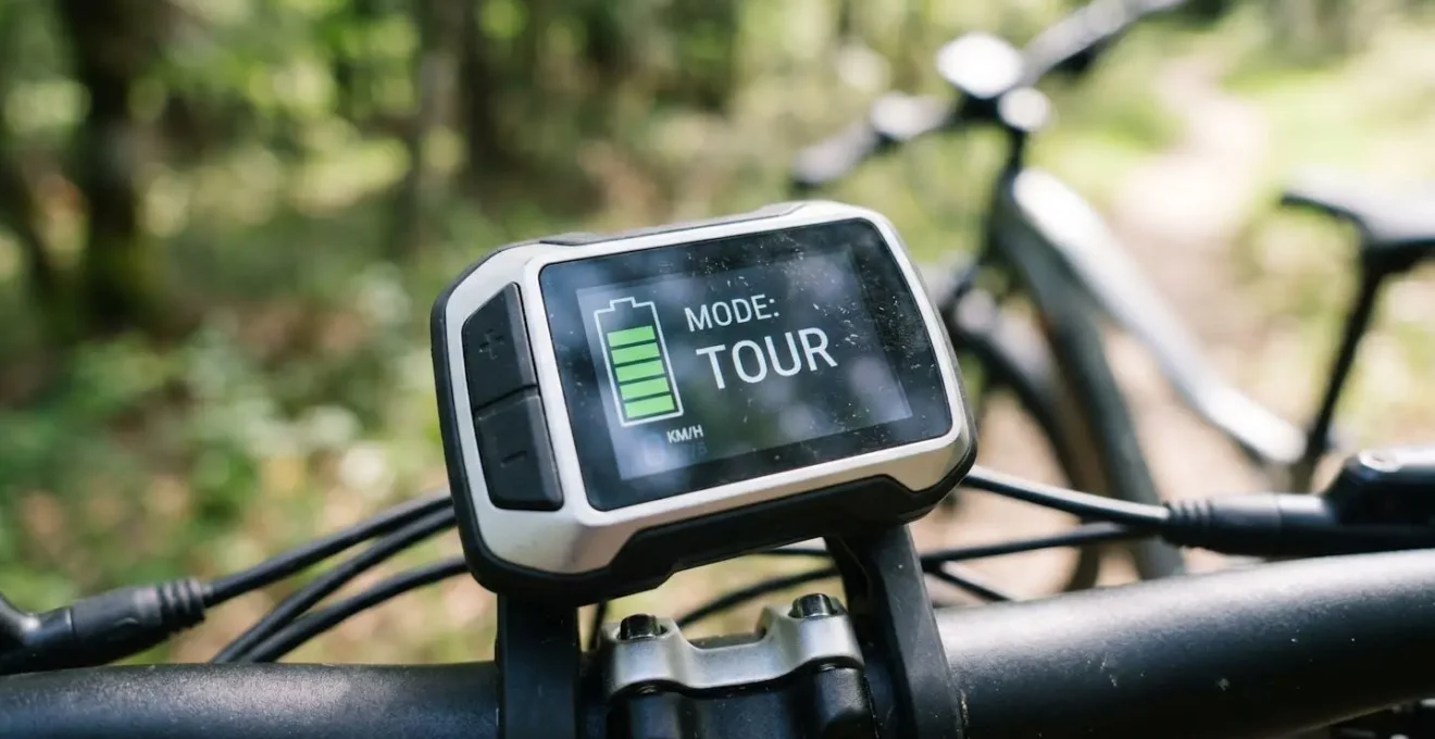 Gros plan sur l'écran d'un vélo électrique affichant le niveau de batterie sous forme de barres et l'indicateur du mode d'assistance actif, avec un arrière-plan flouté de nature et de vélo